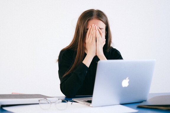 Eine Person, die sich die Hände vor das Gesicht hält und vor sich einen Laptop stehen hat.