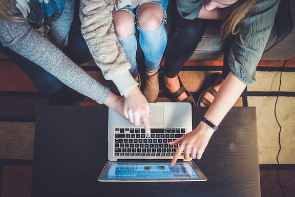 Drei Personen, die sitzend mit ihren Fingern auf den Bildschirm eines Laptops zeigen.