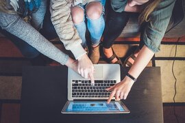 Drei Personen, die sitzend mit ihren Fingern auf den Bildschirm eines Laptops zeigen.