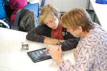Ein Kind und eine erwachsene Person sitzen zusammen an einem Tisch, auf dem ein Tablet liegt.