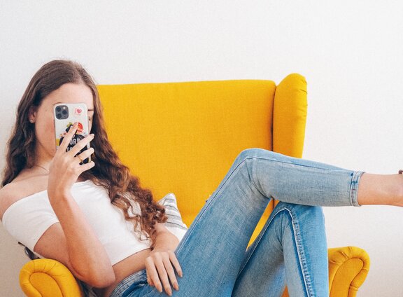 Eine Person, die auf einem Sessel sitzt und ein Smartphone vor ihr Gesicht hält.