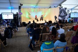 Personen, die auf Stühlen vor einer Bühne sitzen und den Personen auf der Bühne zuhören.
