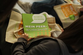 Eine sitzende Person, dauf dessen Schoß ein Heft mit dem Titel "Mein Peer-Workshop" liegt.