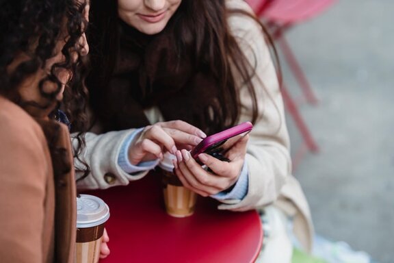 Zwei Personen, die an einem Tisch stehen und von denen eine, ein Smartphone in den Händen hält.