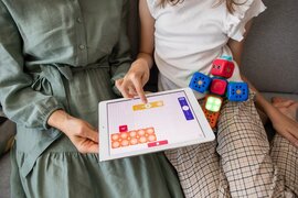 Eine erwacchsene Person und ein Kind, die ein Tablet vor sich liegen haben, auf welches das Kind mit den Fingern zeigt.