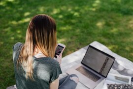 Eine Person, vor der ein Laptop steht und die ein Smartphone in den Händen hält.