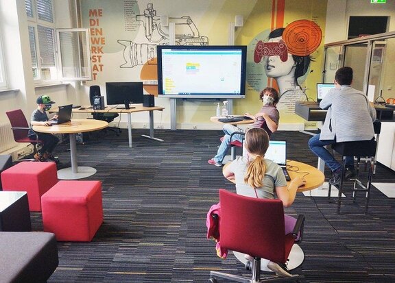 Computerspielschule Stuttgart: Die Lehrperson dreht sich um und blickt zu seinen drei Schüler:innen, welche jeweils an einem Tisch mit Laptop sitzen.