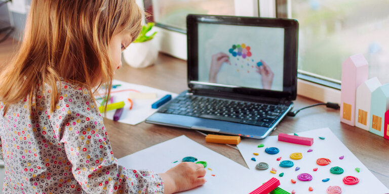 Blondes Mädchen von hinten sitzt an einem Tisch und formt verschiedene Formen aus Knete auf Papier, im Hintergrund ist ein Laptop zu sehen, auf dem die Knet-Anleitung gezeigt wird