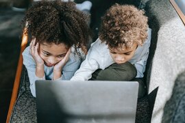 Zwei Kinder, die auf einem Sofa liegen und mit einem Laptop beschäftigt sind.