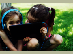 Zwei Kinder, die ein Tablet vor ihre Gesichter halten und auf einer Wiese sitzen.