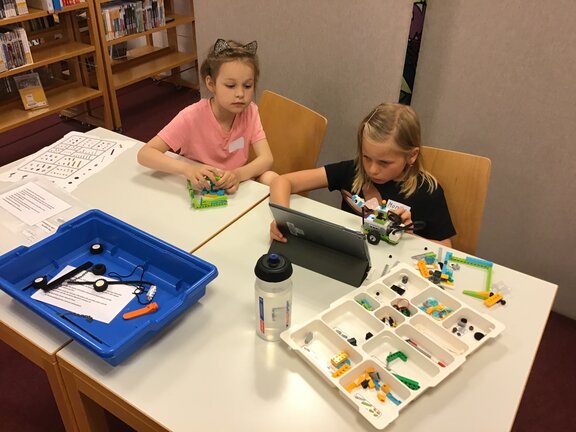 Zwei Kinder, die an einem Tisch sitzen, auf dem ein Tablet und Lego stehen.