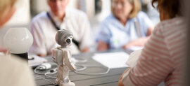 Ein kleiner Roboter steht auf einem Tisch. Im Hintergrund erkennt man verschwommen vier Senior*innen, die um den Roboter herum sitzen und ihn bestaunen. 