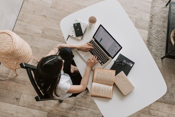 Eine Person, die an einem Schreibtisch sitzt, auf der ein Laptop steht und mehrere Bücher liegen.