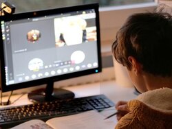 Ein junger Mensch, der vor einem PC sitzt, ein Arbeitsheft vor sich auf dem Schreibtisch liegen hat und einen Stift in der Hand hält.