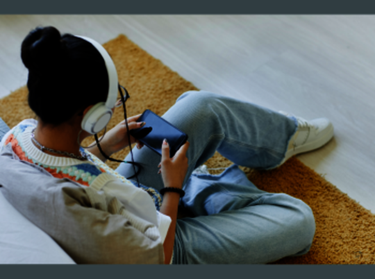 Eine Person, die auf dem Boden sitzt, ein Smartphone in den Händen hält und Kopfhörer aufhat.