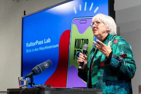 Die Bundesministerien Claudia Roth steht rechts vor einem Bildschirm, auf dem eine Grafik zu dem KulturPass  sichtbar ist. 