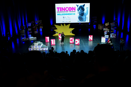 Eine Halle, in der Zuschauer:innen auf eine Bühne schauen, auf der eine Person mit Mikrofon steht.