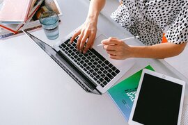 Eine Person, die am Schreibtisch sitzt und einen Laptop und ein Tablet vor sich auf dem Tisch liegen hat.