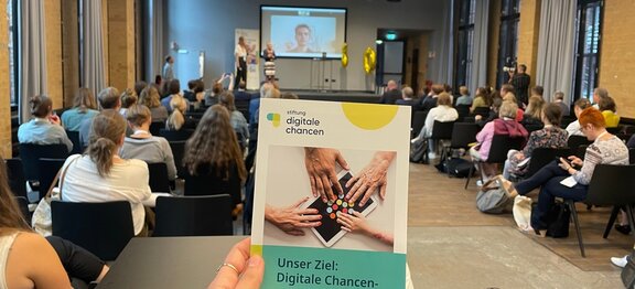 Im Vordergrund wird eine Broschüre von der Stiftung Digitale Chancen gehalten, im Hintergrund ist eine Veranstaltung zu sehen, wobei Personen auf der Bühne sehen und dein Publikum diesen Personen und ihren Vorträgen zuhört.