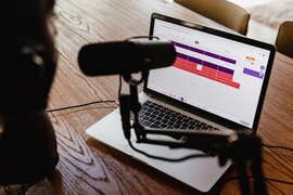 Eine Person, die in ein Mikrofon spricht und einen Laptop vor sich auf einem Tisch stehen hat.