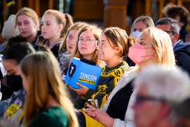 Junge Menschen, die nebeneinander sitzen und Menschen auf einer Bühne zuhören.