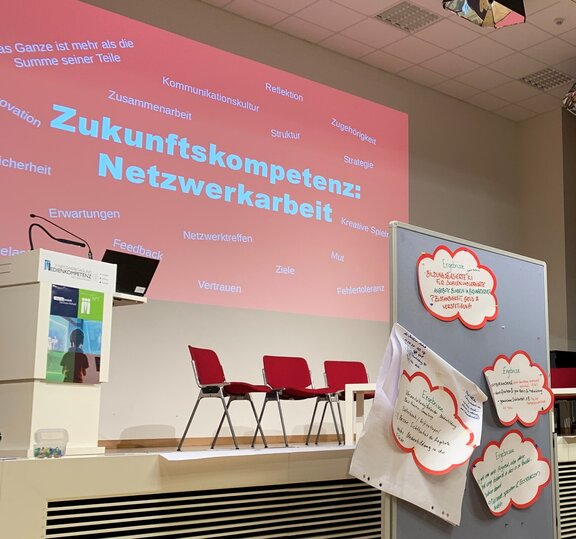 Eine Bühne, auf der Stühle stehen. Vor der Bühne steht eine Pinnwannd, an der Zettel hängen.